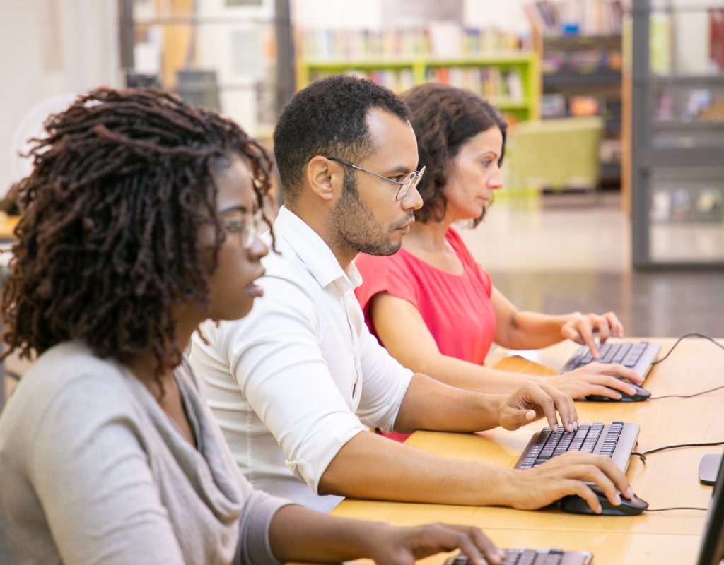 Diverse group of adult students working in computer class. Line of man and women in casual sitting at table, using desktops, typing, looking at monitor. Training center concept