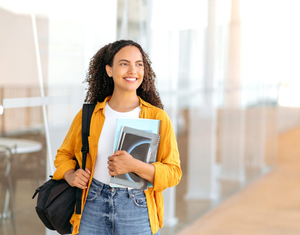 Education concept, female student. Happy lovely brazilian female student, with a backpack, hold books and notebooks in her hand, stand near the university campus, looks away and smile. Copy-space