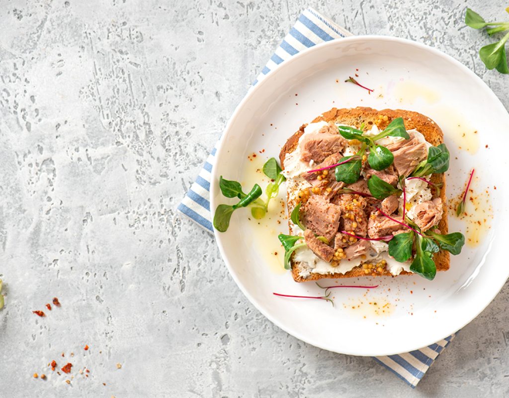 Tasty and healthy breakfast or lunch. Toast with tuna, ricotta cheese, and herbs in a white plate on a gray background top view. Mediterranean style breakfast. Free space for text.