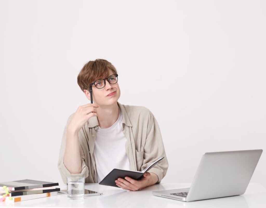 closeup-of-pensive-handsone-young-man-student-wears-beige-shirt-thinking-and-writing-in-notebook-at-the-table-with-laptop-computer-isolated-over-white-wall