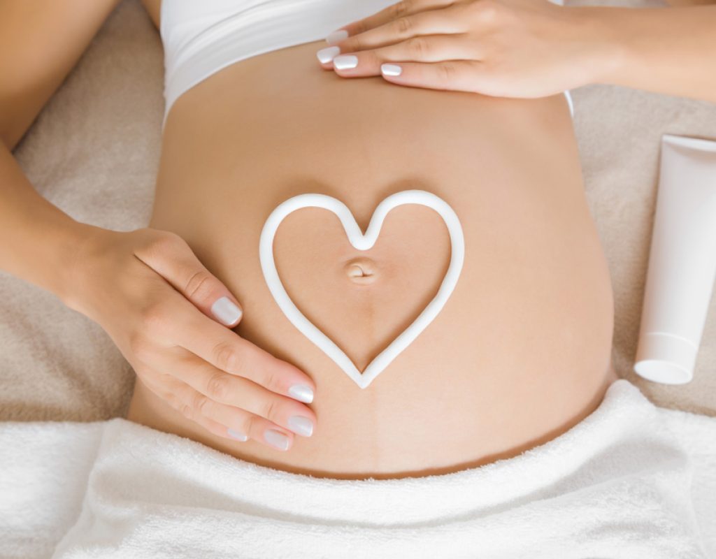 Young woman lying down on bed. Gentle hands touch.  White heart shape created from cream on naked big belly. Care about perfect and smooth skin in pregnancy time. Close up.